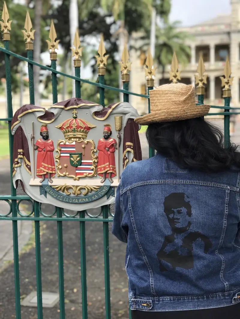 A woman stands before the royal gates bearing the Hawaiian coat of arms.